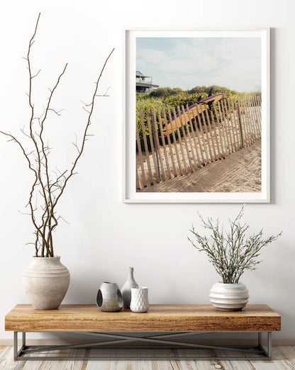 Ditch Plains Surfboard art framed above a wooden table with vases, greenery, and a branch in a minimalist room.