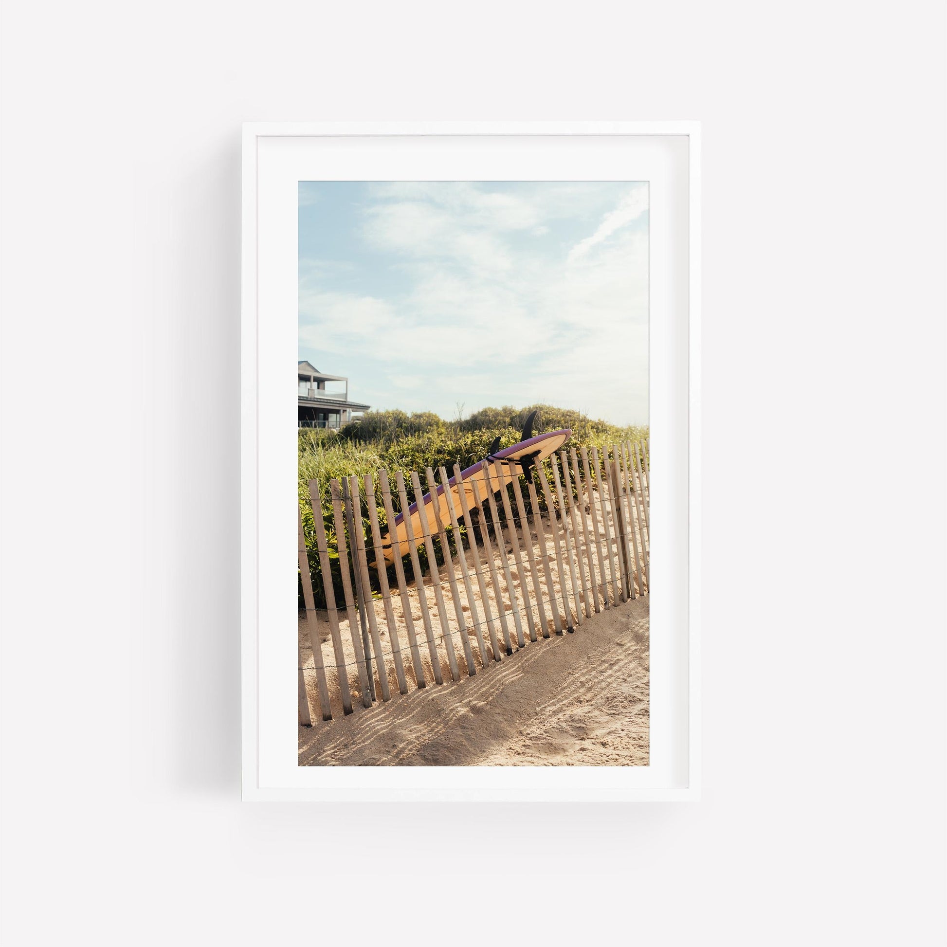 A wooden fence lines a sandy shore with the Ditch Plains Surfboard and greenery beneath a partly cloudy sky.