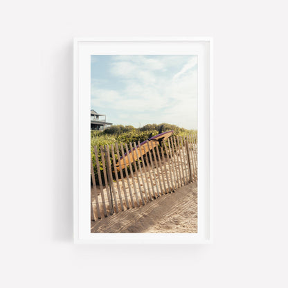 A wooden fence lines a sandy shore with the Ditch Plains Surfboard and greenery beneath a partly cloudy sky.