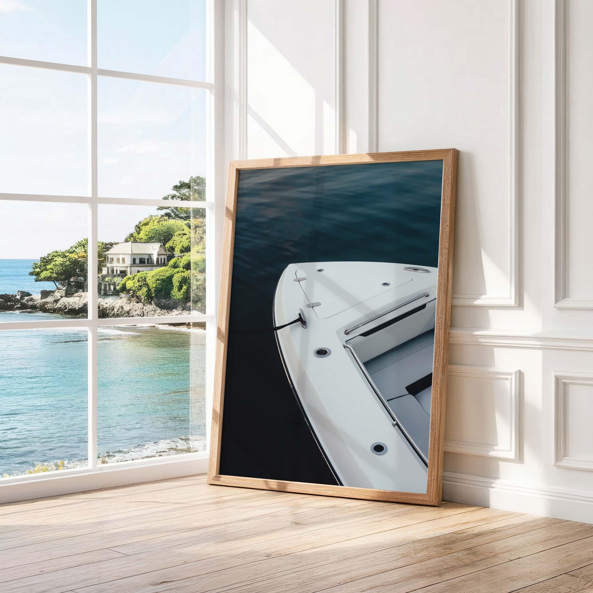 Blue Hour Mooring“ framed photo of a yacht’s bow leans against a white wall by an ocean-view window.
