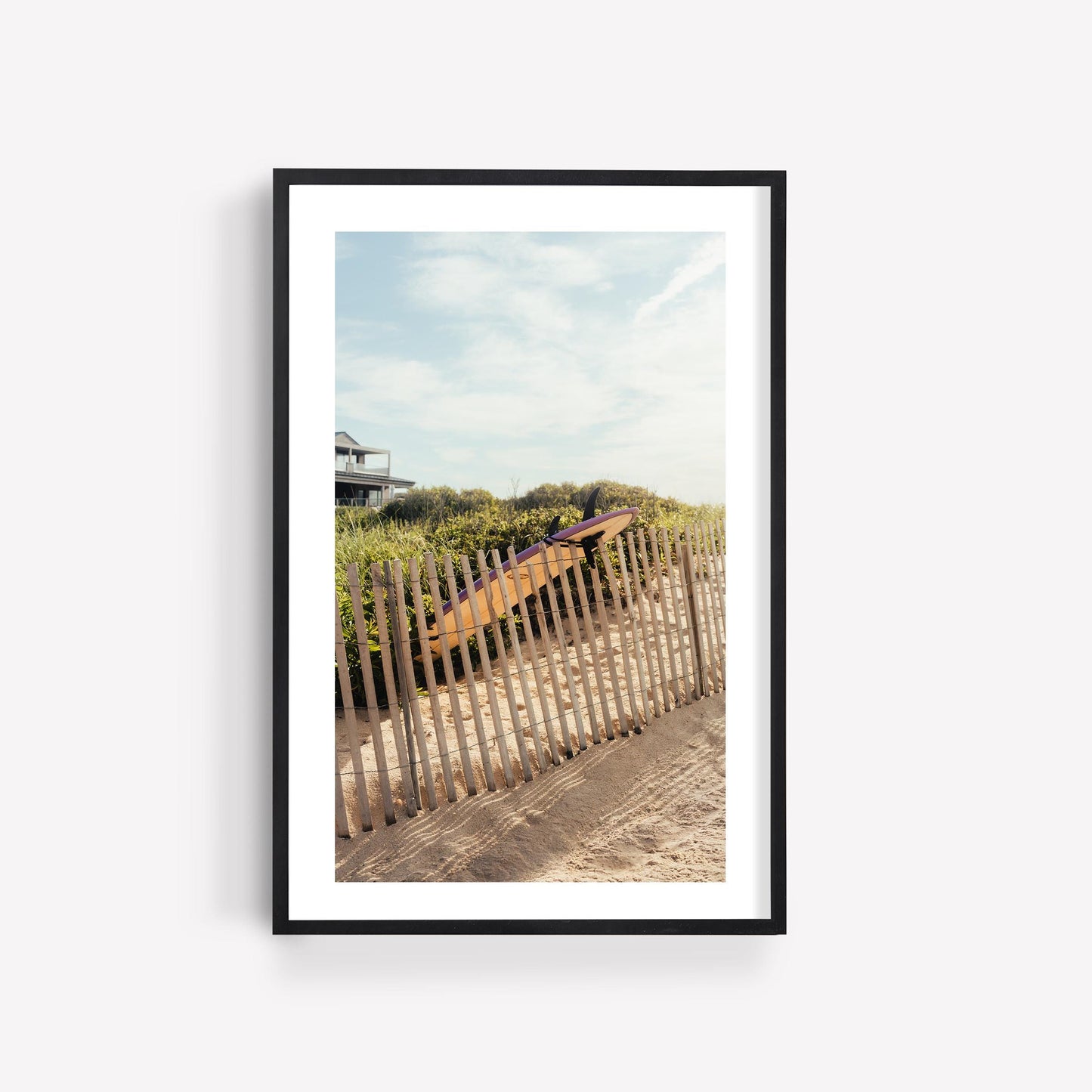 Framed photo: Ditch Plains Surfboard by a wooden fence on a sandy beach.