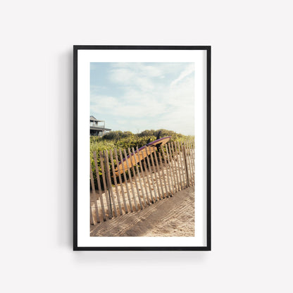 Framed photo: Ditch Plains Surfboard by a wooden fence on a sandy beach.