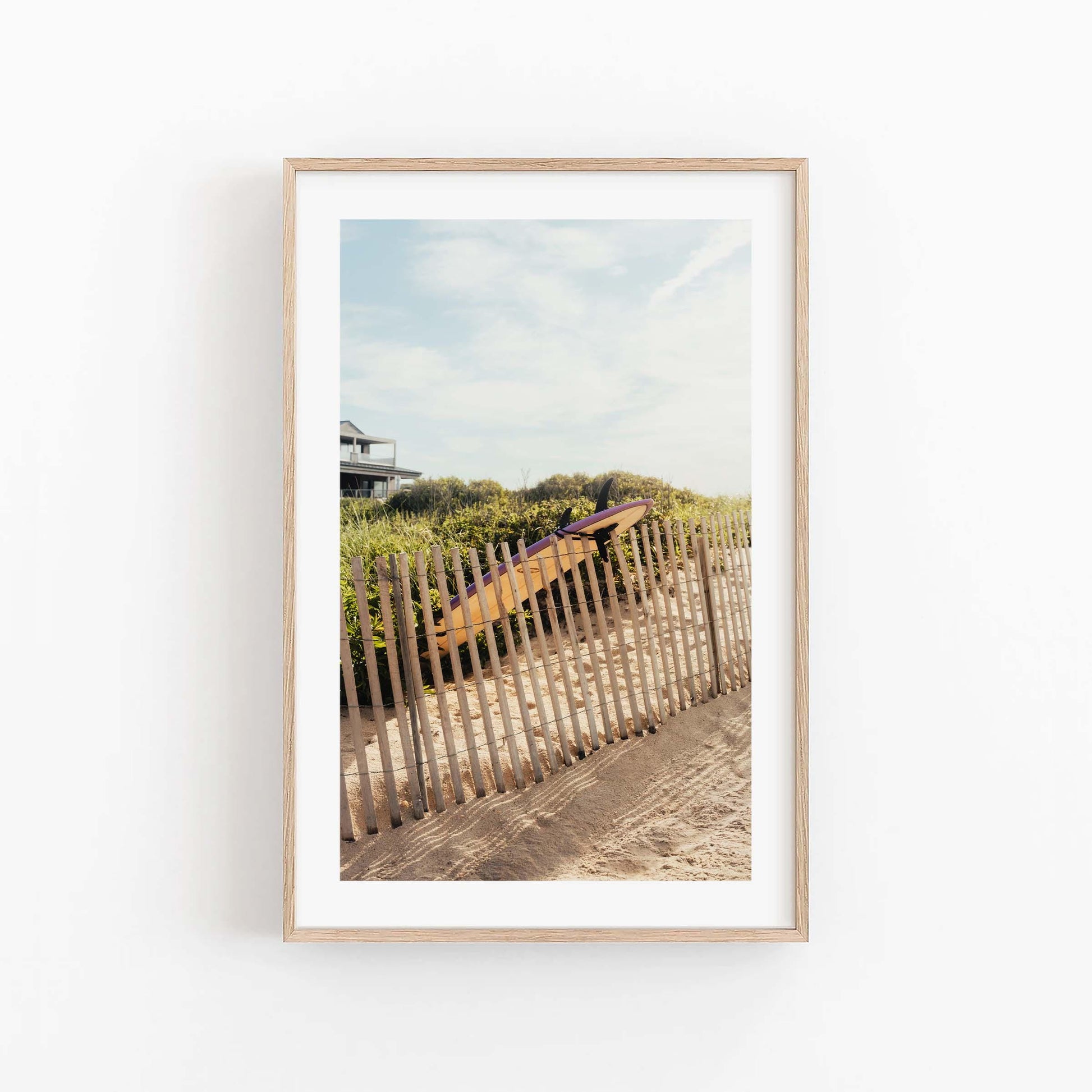 Framed photo of Ditch Plains Surfboard on sandy path with fence, beach grass, house, and blue sky in background.
