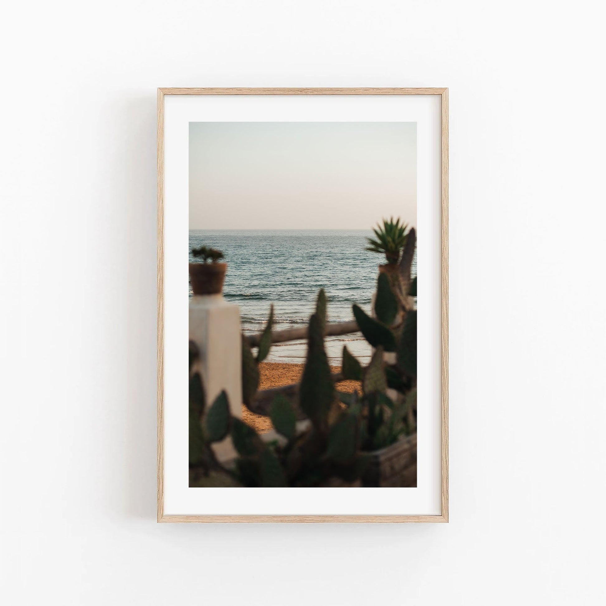 The In the Distance framed photo of a beach scene with cacti and waves hangs on a white wall.