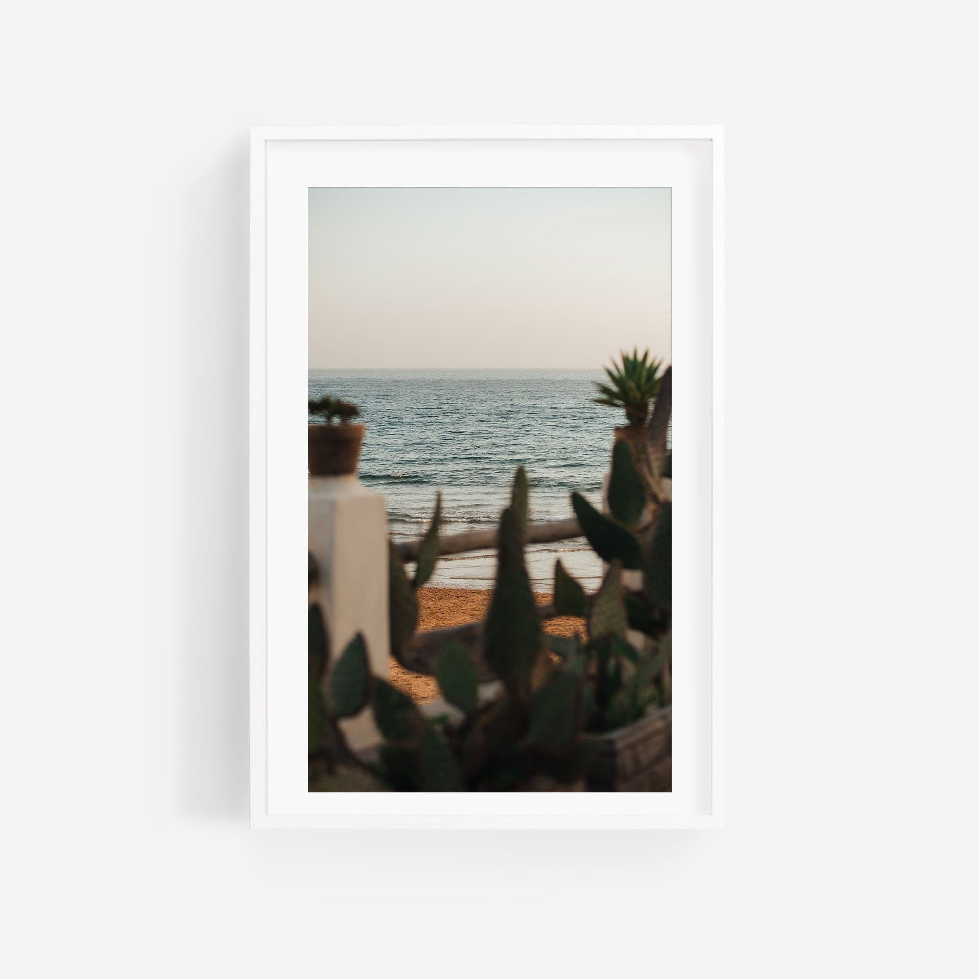 In the Distance framed photo captures a tranquil beach scene with calm waves and blurred foreground plants.