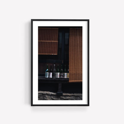 Framed photo titled Japanese Serenity - Set of 3 features sake bottles lined up under bamboo blinds against a dark background.