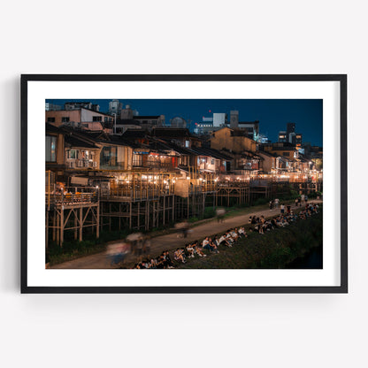 Framed photo of Kamo River at night, featuring illuminated buildings and people gathered along the riverbank.