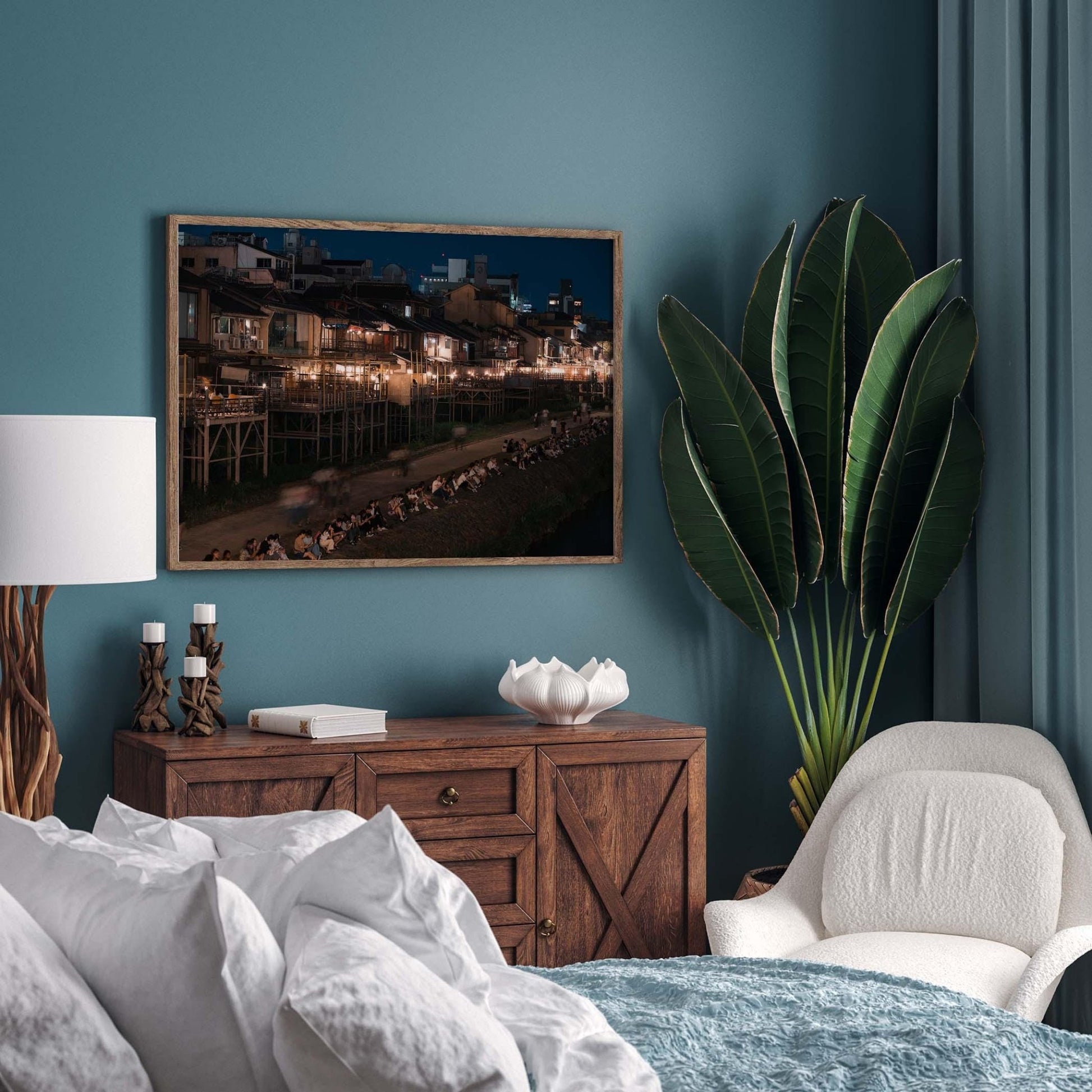 Cozy bedroom featuring the Kamo River framed cityscape photo, a green plant, wooden dresser, and white armchair against a teal wall.