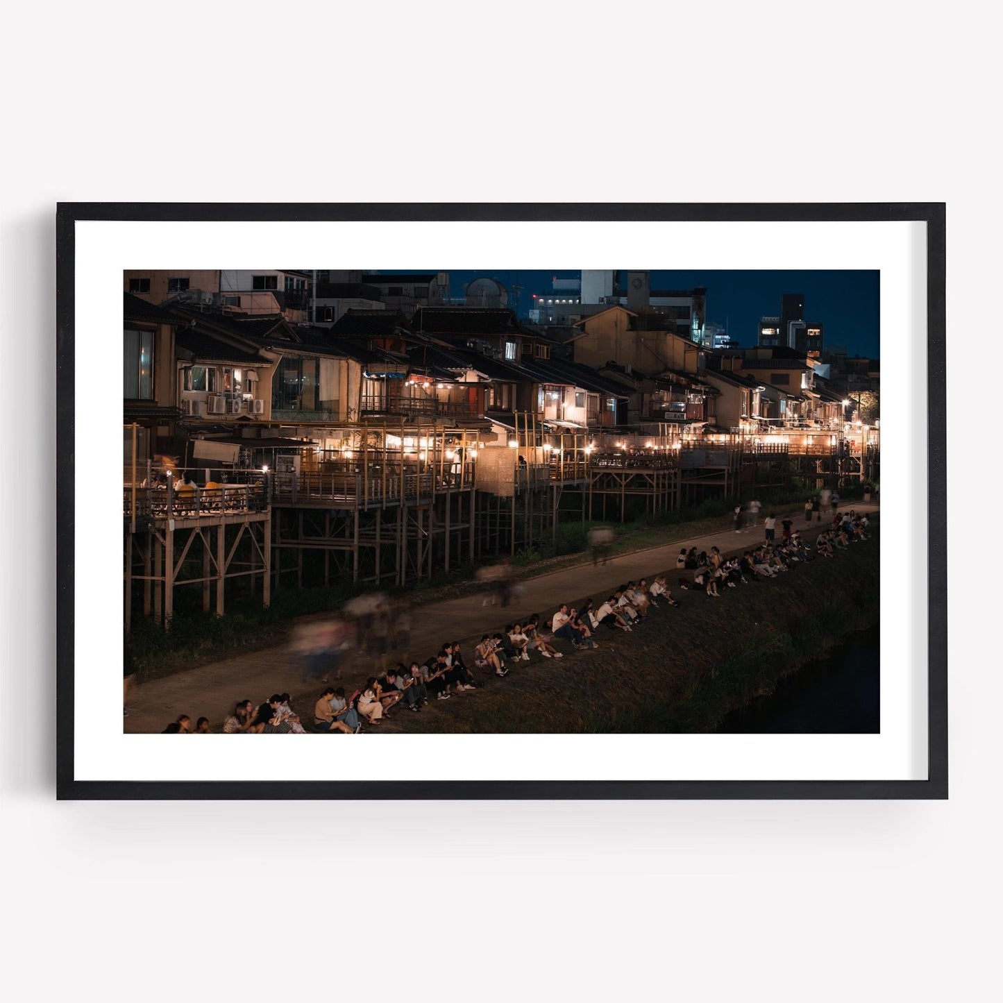 Framed photo titled Kamo River, featuring people sitting by a riverbank at night with illuminated buildings in the background.