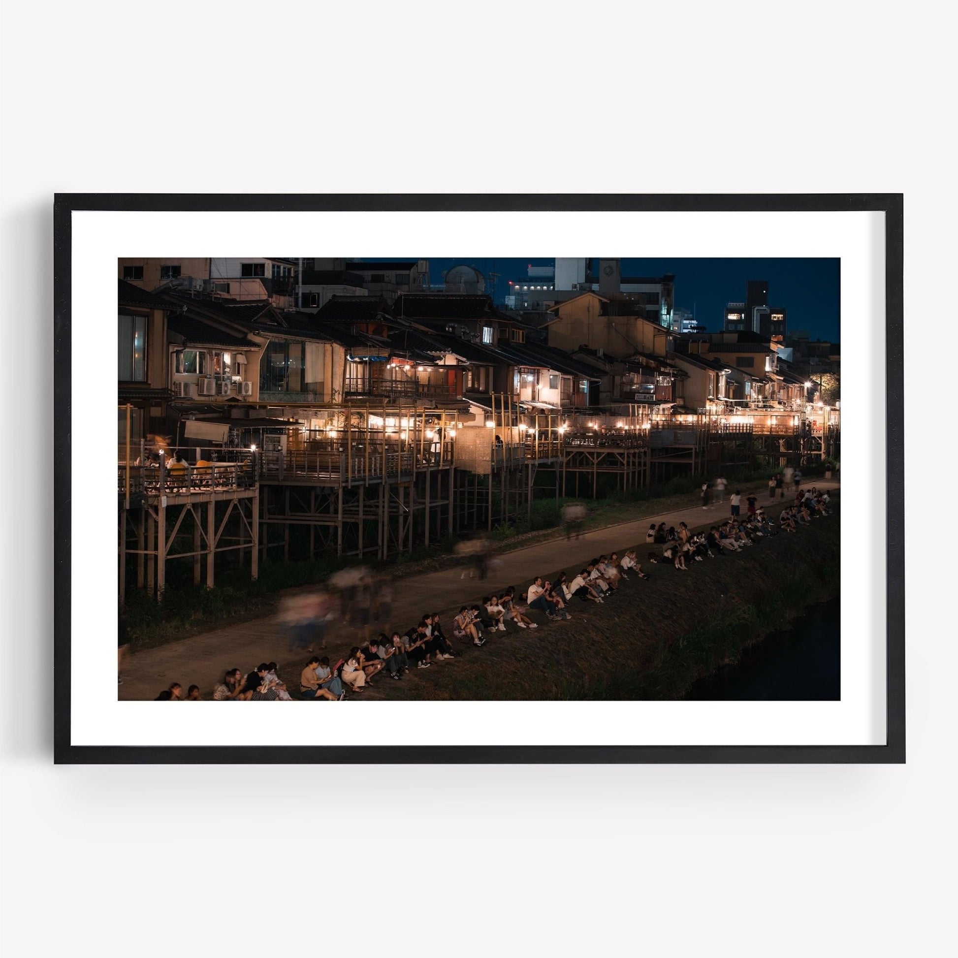 Framed photo titled Kamo River, featuring people sitting by a riverbank at night with illuminated buildings in the background.