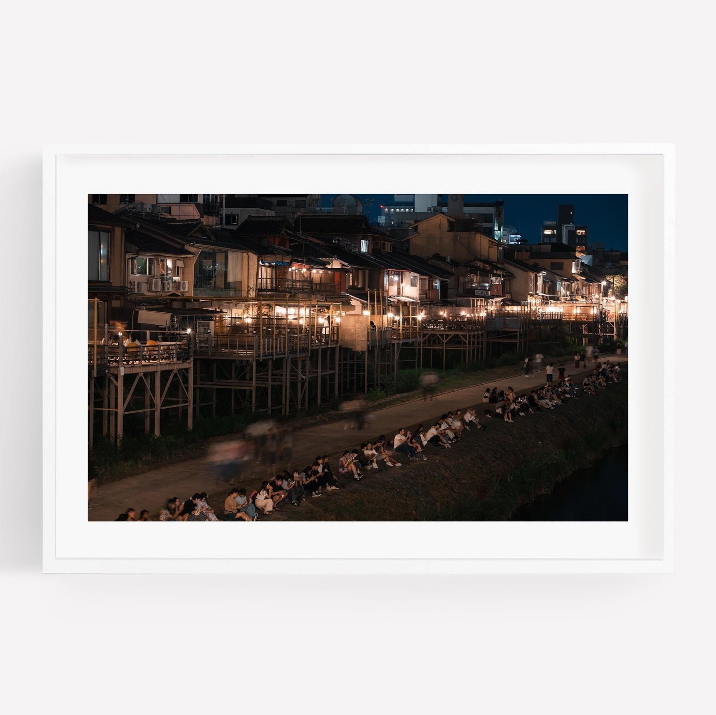 Night scene of Kamo River with people sitting along the riverbank and lit-up buildings in the background.