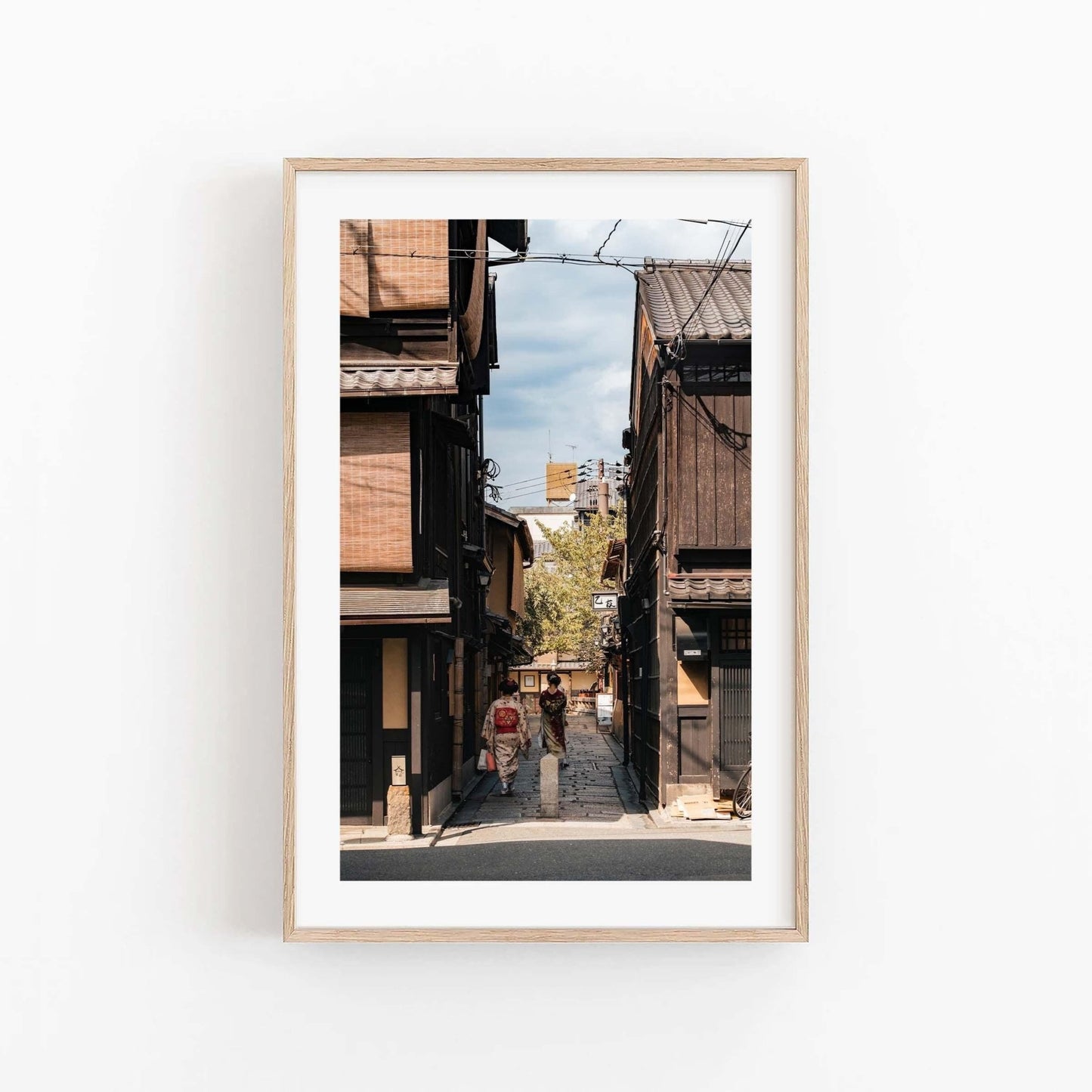 The framed photo Kimonos in Gion, featuring a narrow street with traditional wooden houses and pedestrians, is displayed on a white wall.