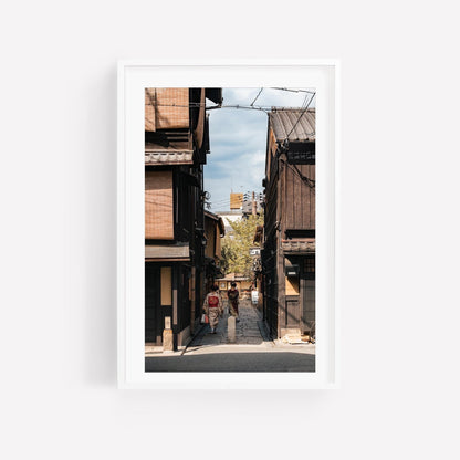 A framed photo titled Kimonos in Gion showcases people walking along a narrow street lined with traditional wooden buildings beneath a blue sky.