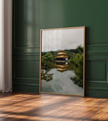 A framed photo of the Kinkaku-ji Temple by a lake, leaning against a dark green paneled wall on a wooden floor.