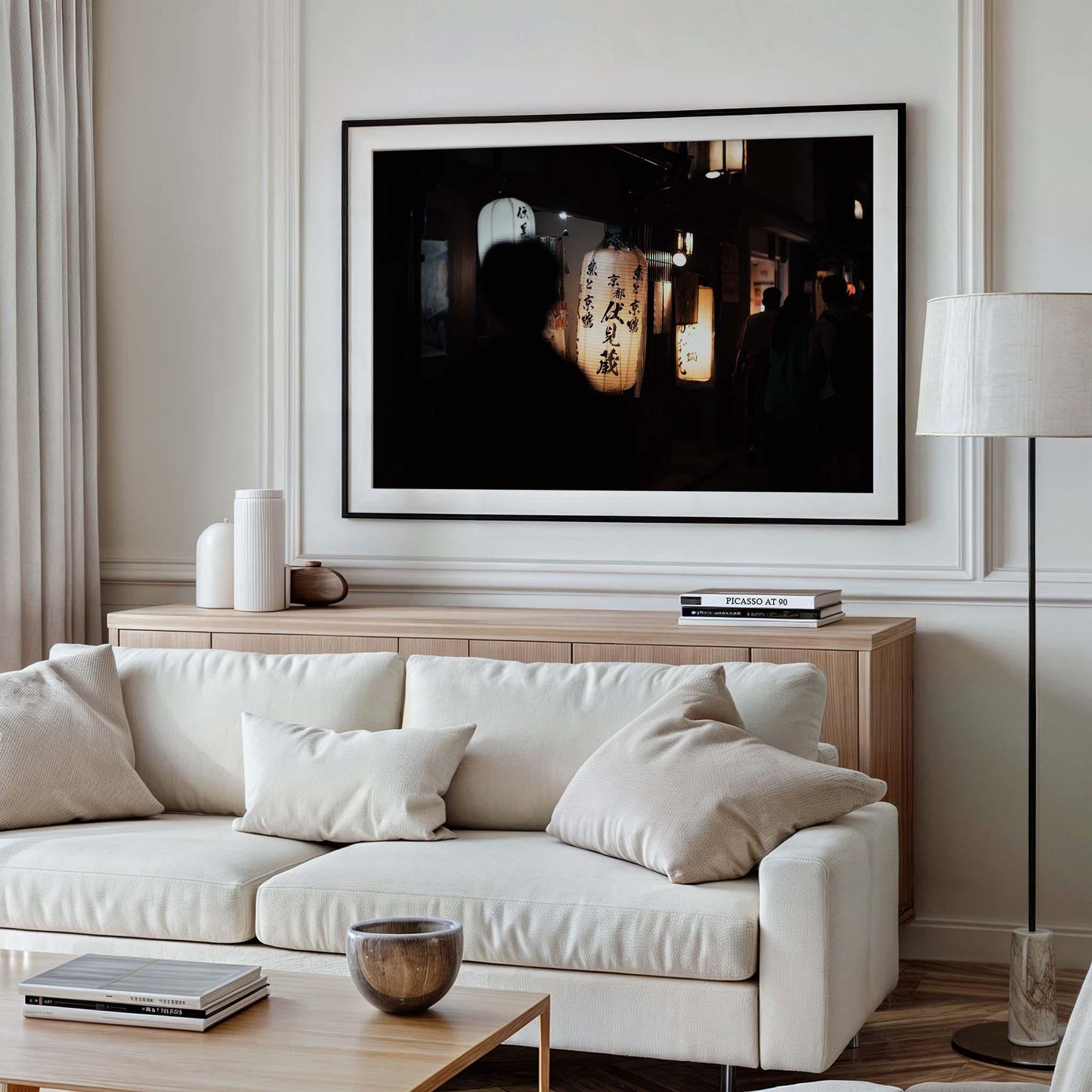 A minimalist living room features a white sofa, pillows, and a lamp, complemented by the framed wall art Kyoto At Night, depicting lanterns. Neutral tones prevail throughout the space.