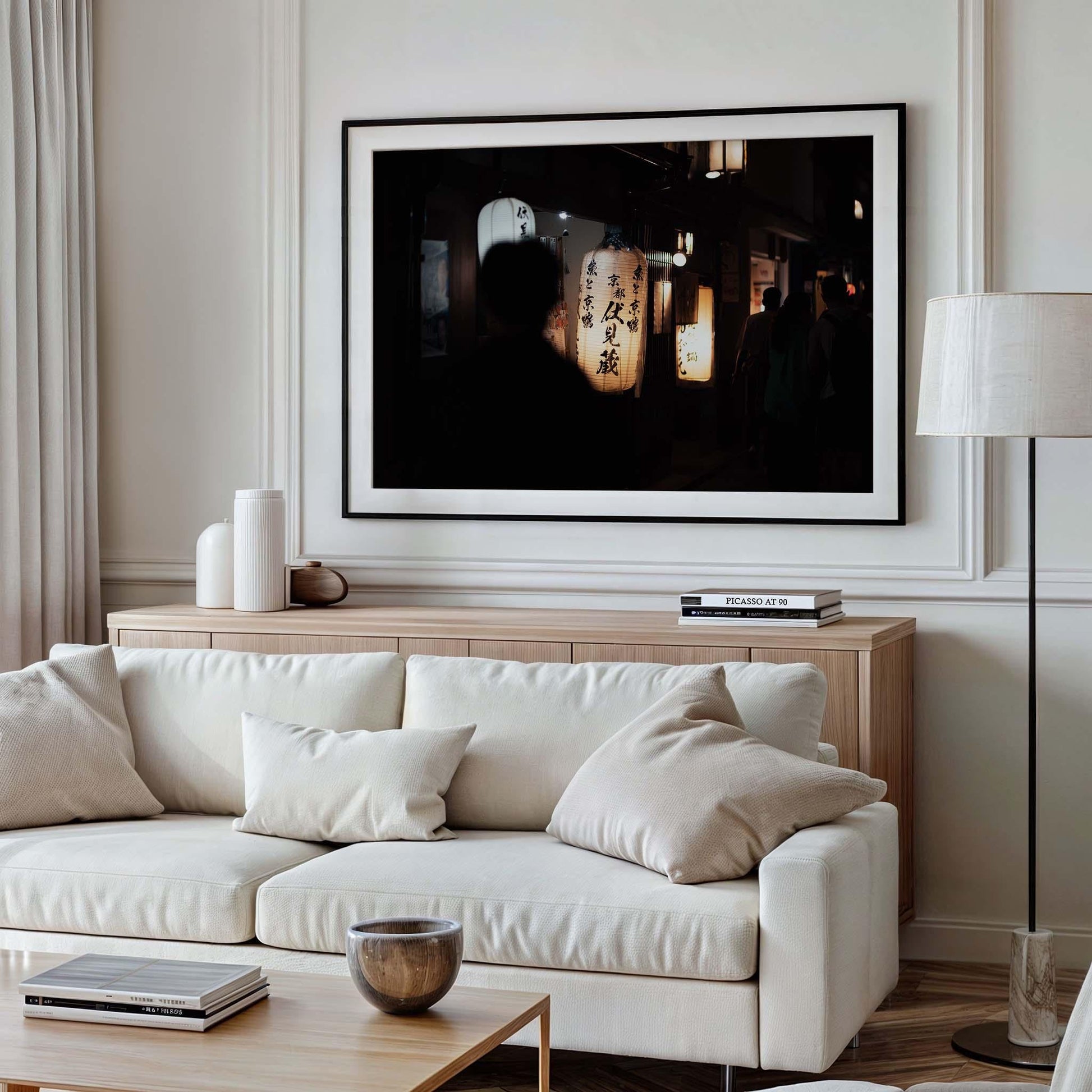 A minimalist living room features a white sofa, pillows, and a lamp, complemented by the framed wall art Kyoto At Night, depicting lanterns. Neutral tones prevail throughout the space.