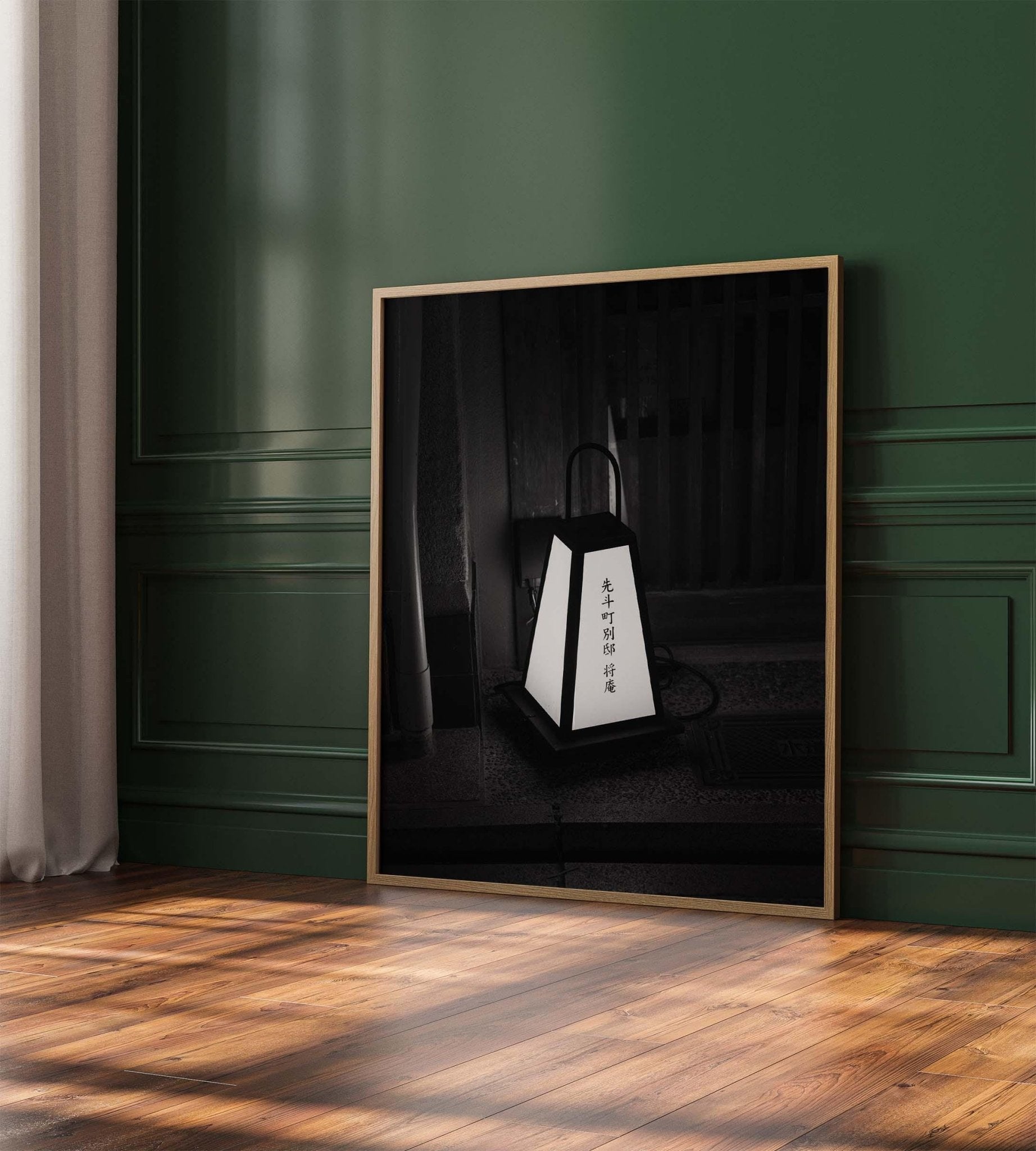 A black and white photo, named Kyoto Lantern I, depicting a lantern with Asian script is set against a green wall on a wooden floor.