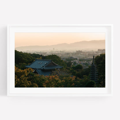 Kyoto Sunset Skyline I: A traditional Japanese building surrounded by trees, set against a backdrop of a cityscape and mountains at sunset.