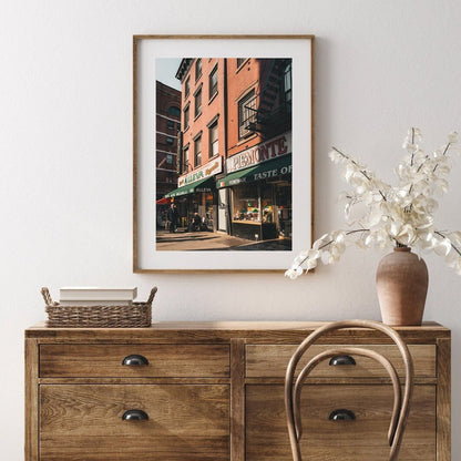 Little Italy city street scene framed photo hangs above wooden dresser with a basket and vase.