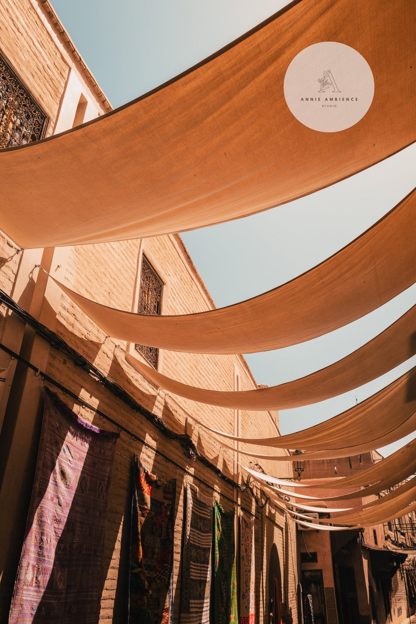 Marrakesh Souk II: Vibrant fabric canopies and colorful rugs adorn brick walls under a clear blue sky.