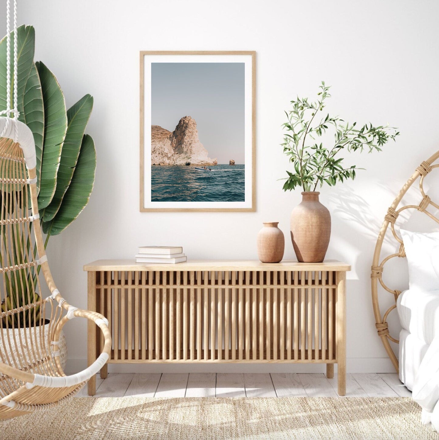 A minimalist room features Mediterranean Adventure art, a wooden cabinet, greenery, with hanging and circular chairs nearby.