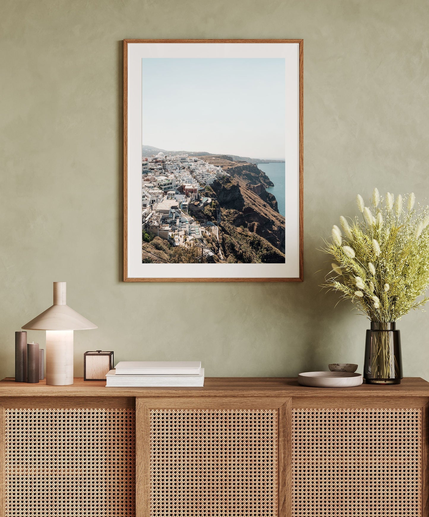 Perched, a framed coastal city photo, hangs above a rattan cabinet with a lamp, books, and a vase of flowers.
