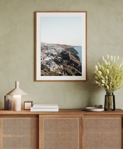 Perched, a framed coastal city photo, hangs above a rattan cabinet with a lamp, books, and a vase of flowers.