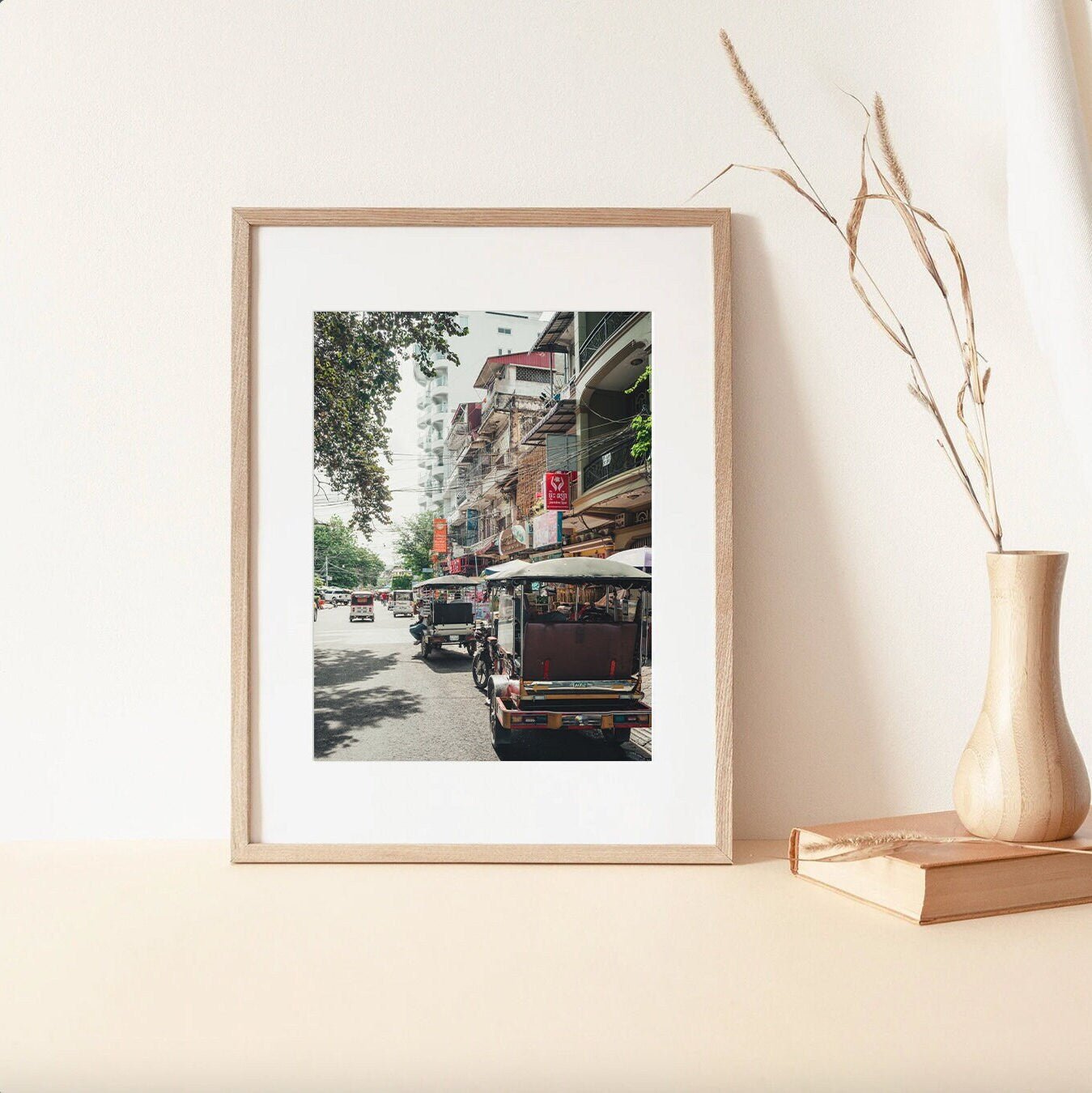 Phnom Penh TukTuk photo with buildings on a light surface, near a tall vase and stacked books.