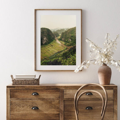 The framed River Valley landscape hangs above a wooden dresser, wicker basket, and vase with white flowers.