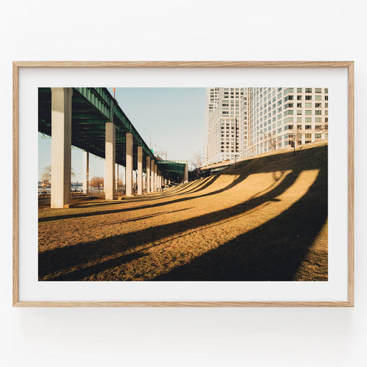 Framed Riverside photo of highway shadows on grass, with tall buildings in the background.