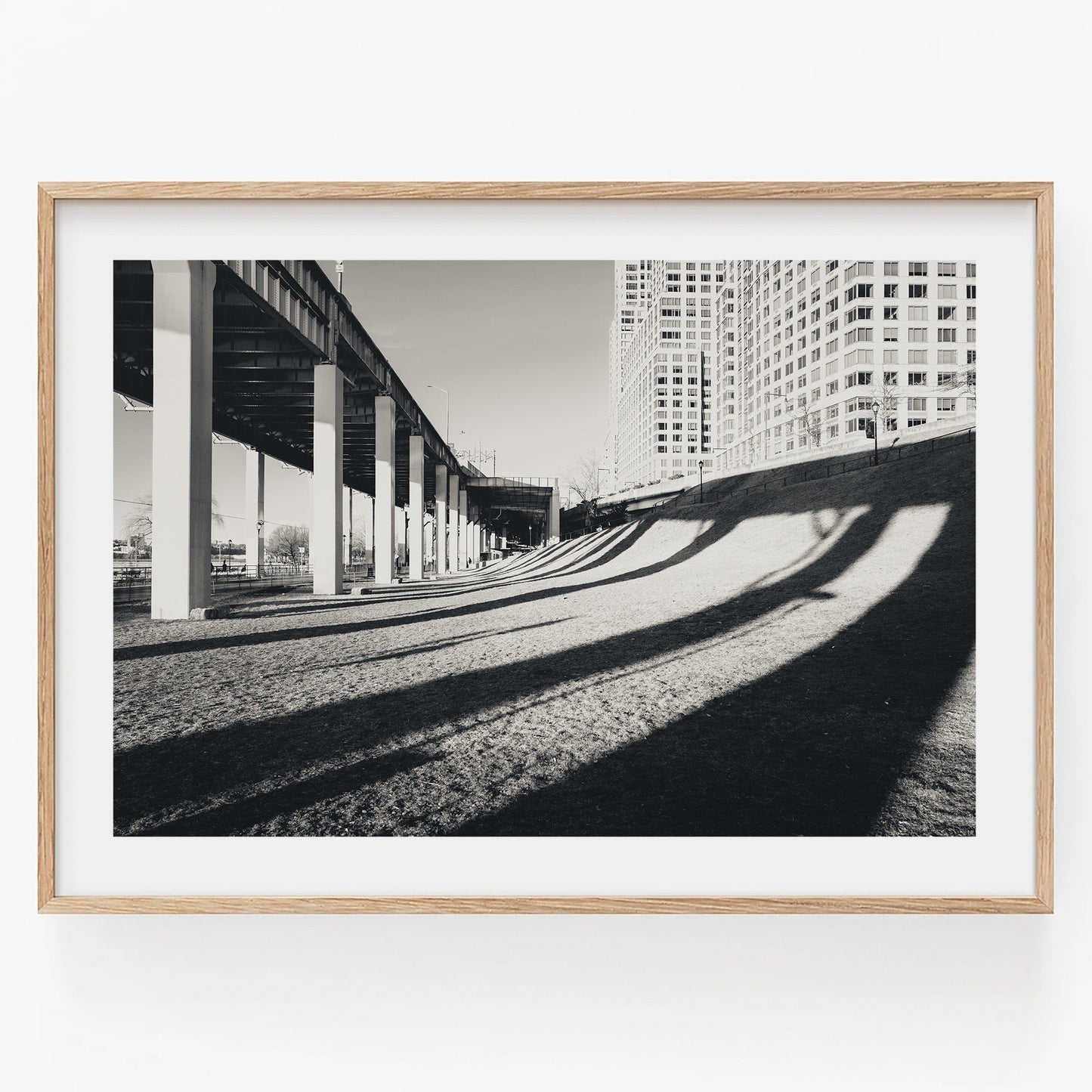 Framed photo Riverside Shadow Black and White of a bridge casting shadows on grass with buildings in the background.