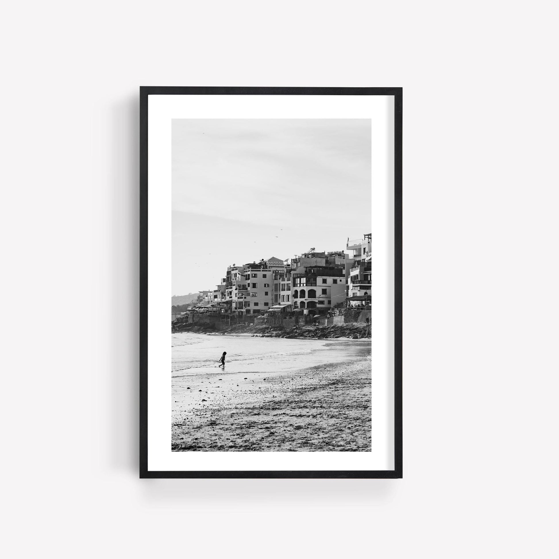 Framed print of Sandy Toes Black and White, a person walking on a beach with buildings in the background.