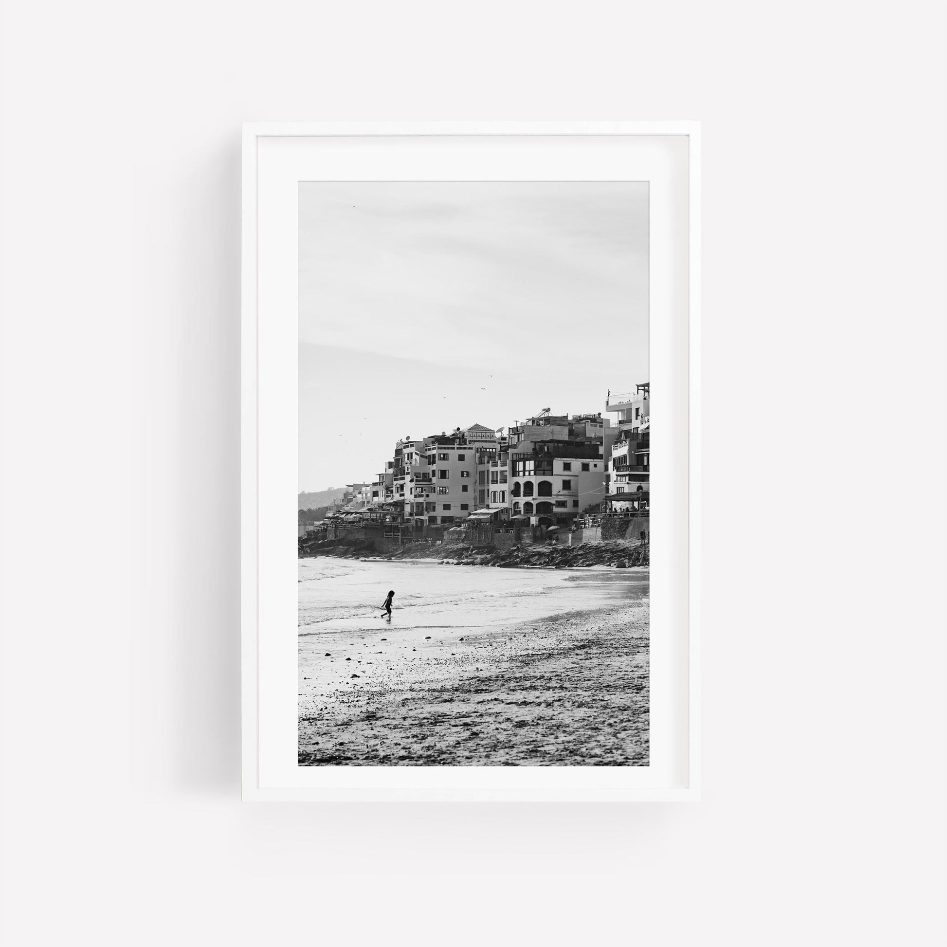 Sandy Toes Black and White: A photo of a person walking on a beach with buildings, framed in white.