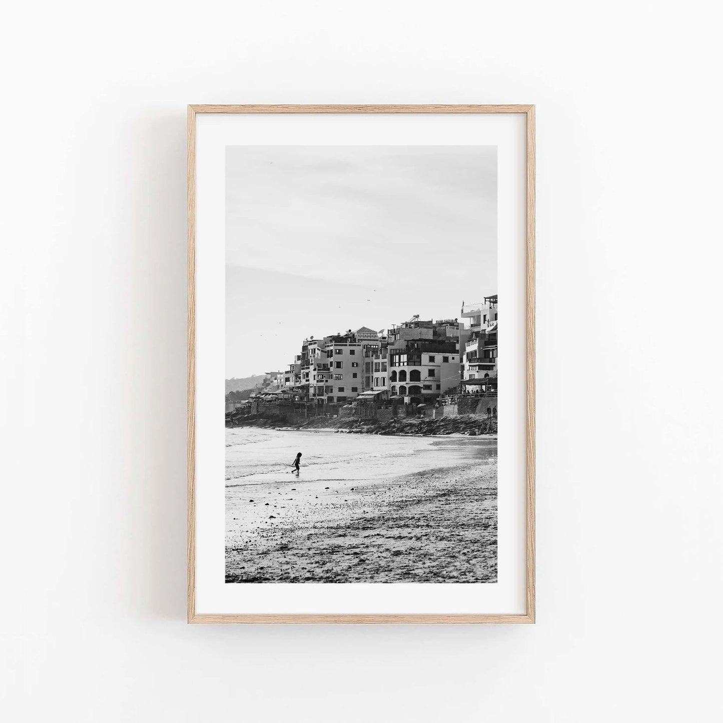 Sandy Toes Black and White: Framed photo of a lone beach walker with hillside buildings in the background.