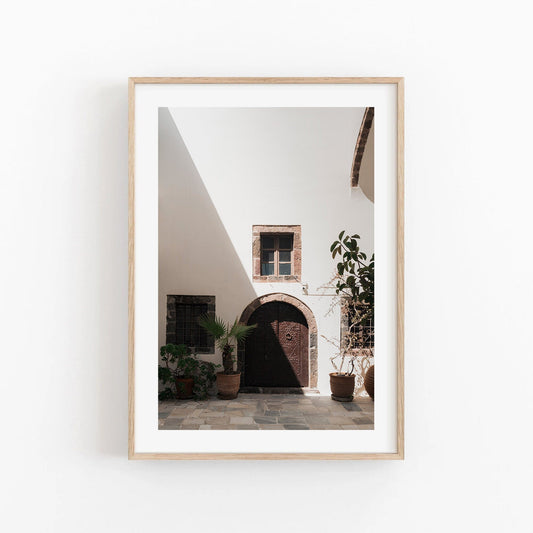 Shadow Slice: Framed photo of a sunny courtyard with an arched wooden door and potted plants.