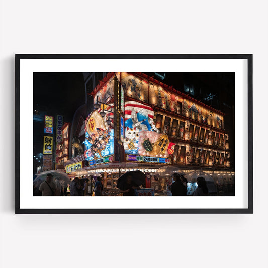 The Shinsekai At Night framed photograph showcases a vibrant, neon-lit building adorned with colorful signage, capturing people with umbrellas below.