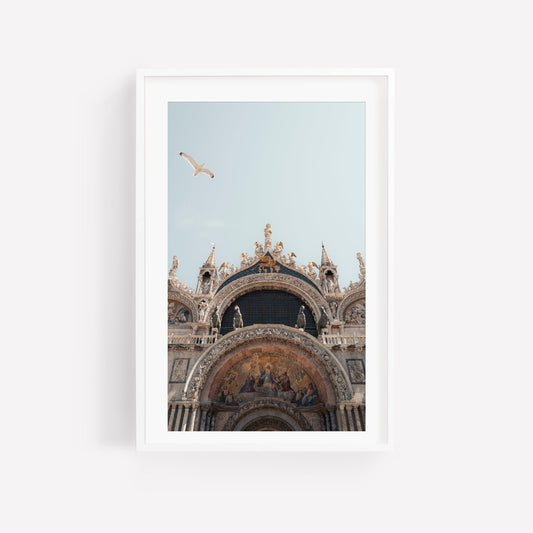 Framed photo of St. Marks Basilica facade with a seagull soaring above against a pale blue sky.