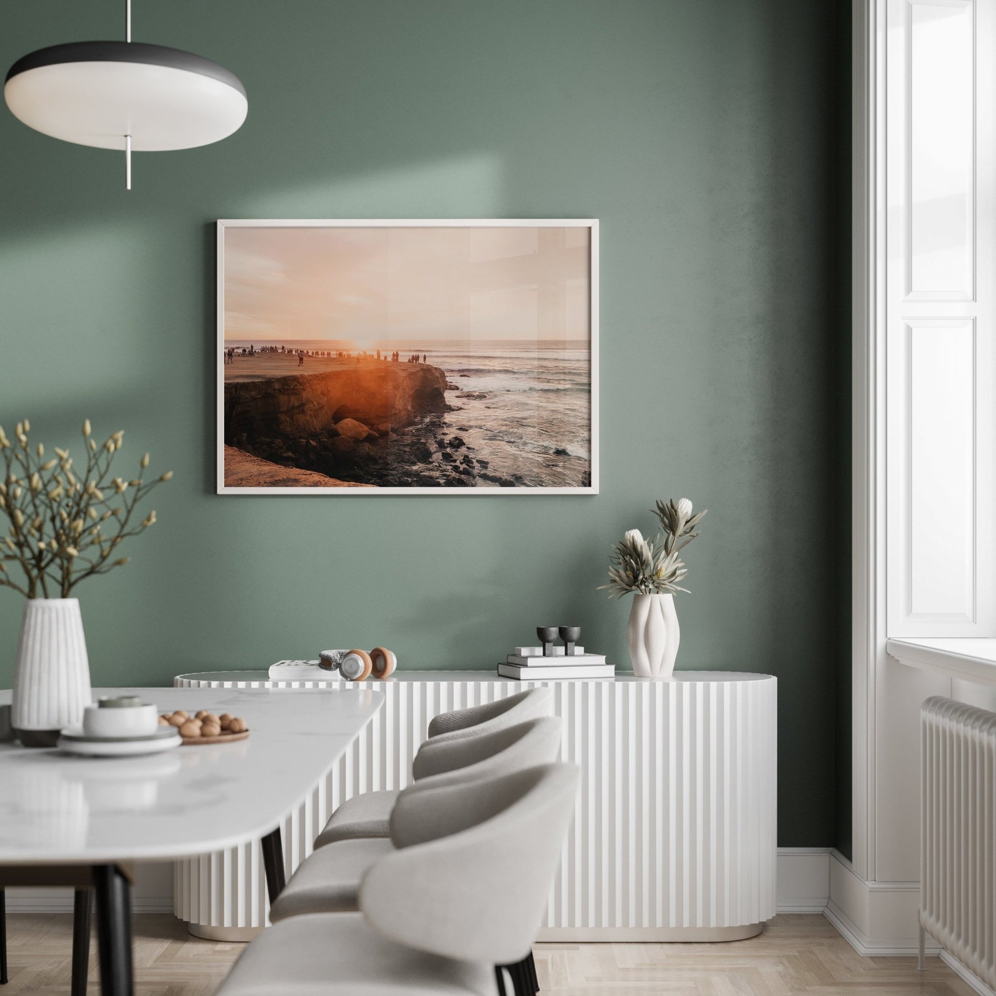 Modern dining room with green walls, framed Sunset Cliffs, white furniture, and minimalistic decor.