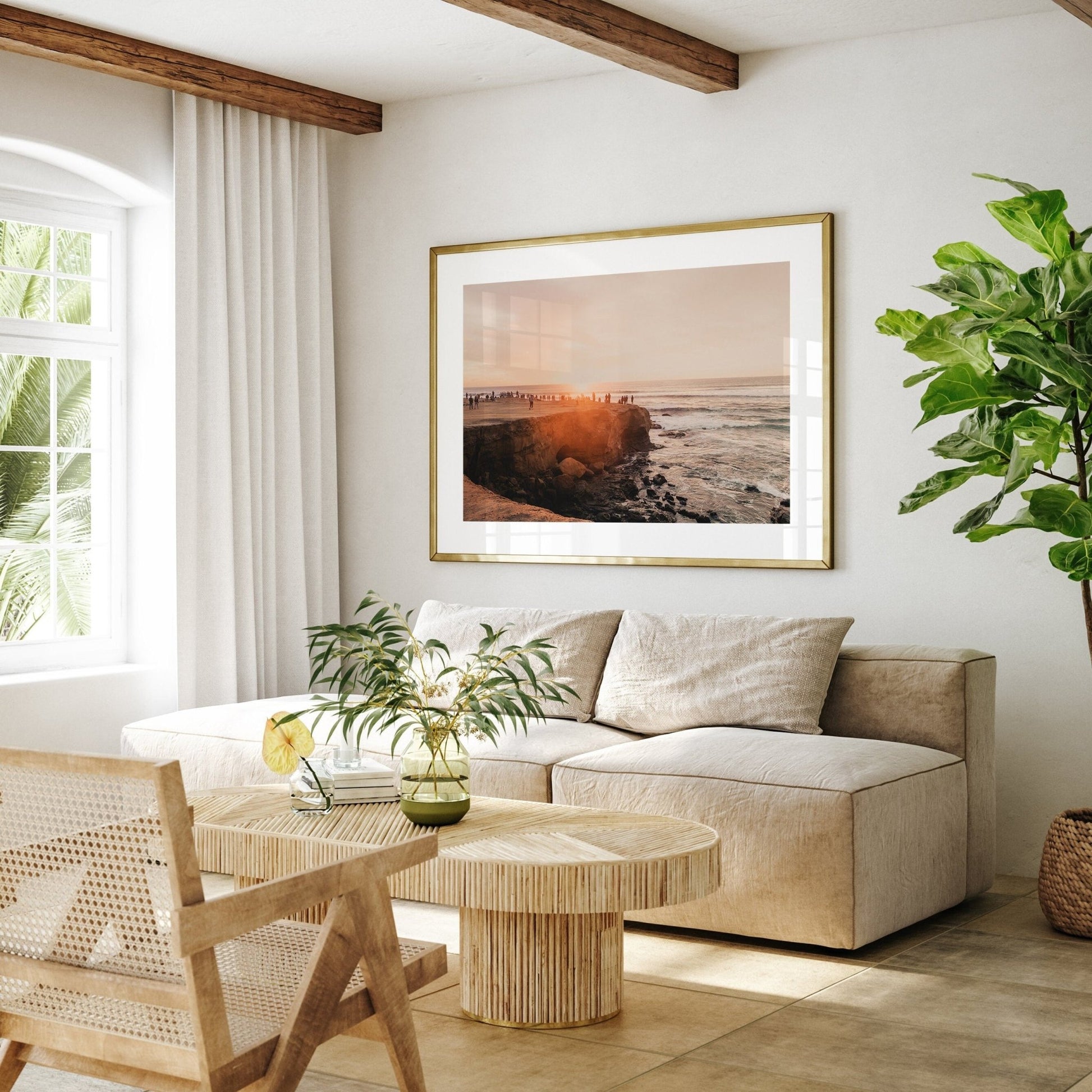 Bright living room with a cozy beige sofa, wooden table, chair, potted plant, and the framed Sunset Cliffs seascape photo.