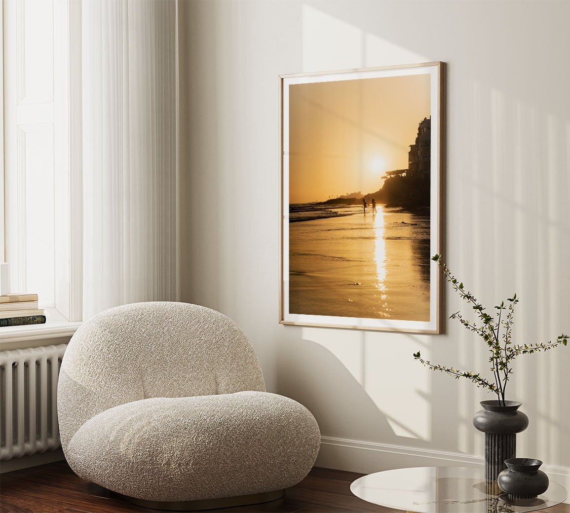 A cozy room with a beige chair, the Taghazout Golden Shores photo on the wall, and a small vase on a table by the window.