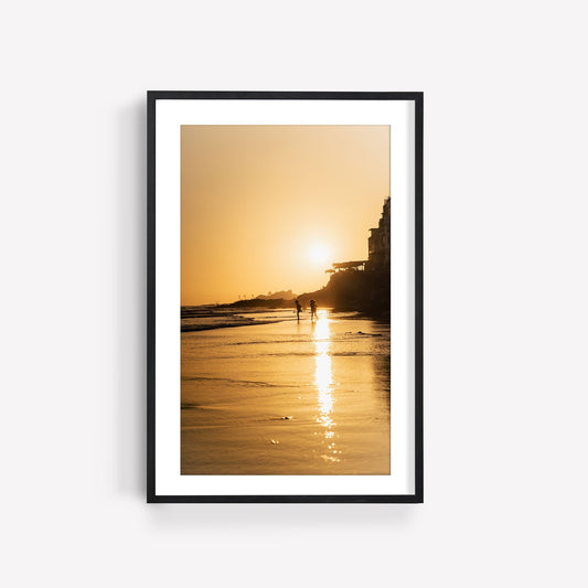 Framed Taghazout Golden Shores photo: sunset beach scene with two people walking along the shoreline, reflected in water.