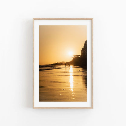 Taghazout Golden Shores: A framed photo of a sunset with two people walking on a beach, orange hues reflecting on water.