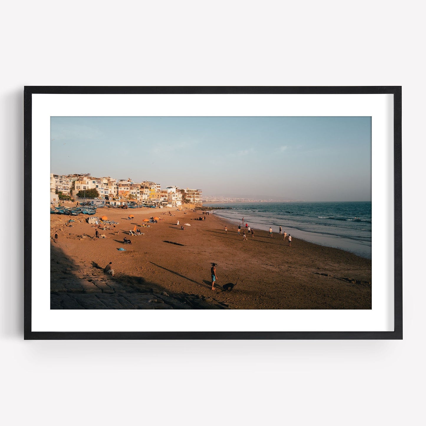 Framed photo of Taghazout I: a sandy beach, people, colorful buildings on the left, and ocean under a clear sky.