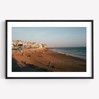 Framed photo of Taghazout I: a sandy beach, people, colorful buildings on the left, and ocean under a clear sky.