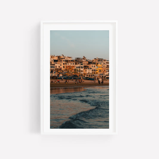 Framed Taghazout Wave photo: lively beach with people, houses, and boats at sunset.