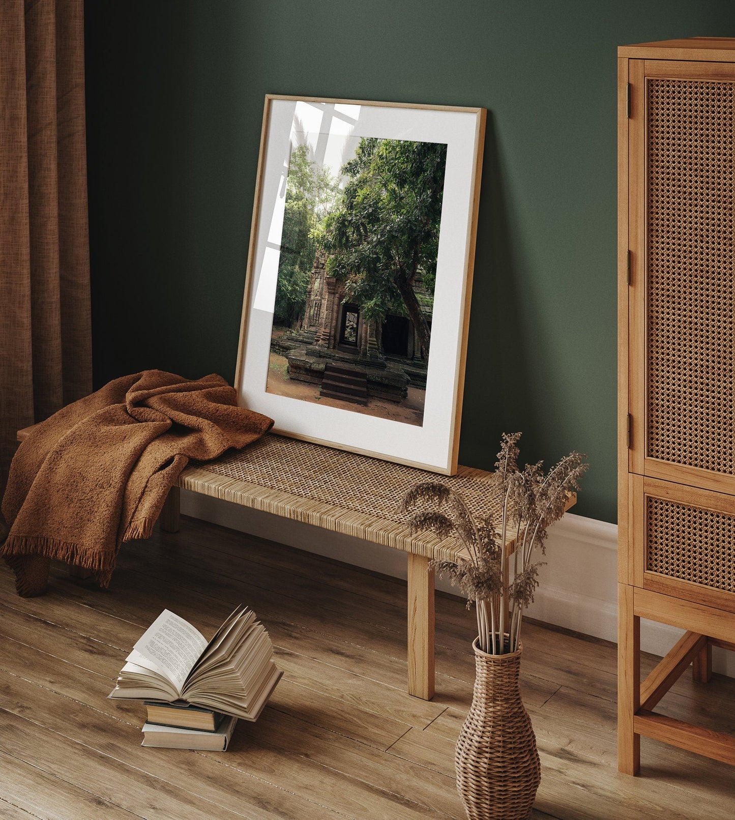 The framed photo Temple II leans on a green wall above a woven bench, beside a book, brown blanket, and dried flowers.