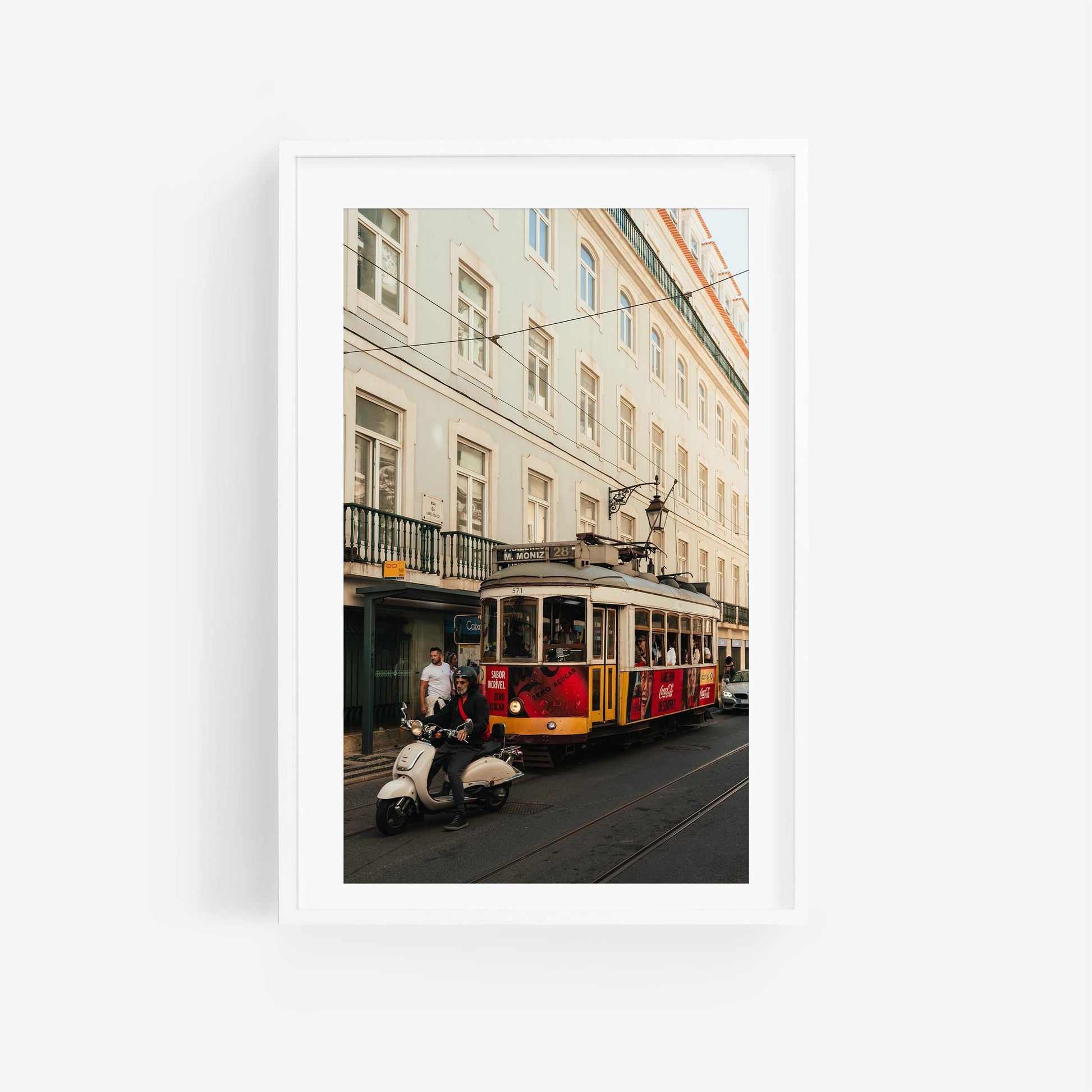 Tram 28 and a scooter rider pass pastel buildings on a city street, all framed in white.