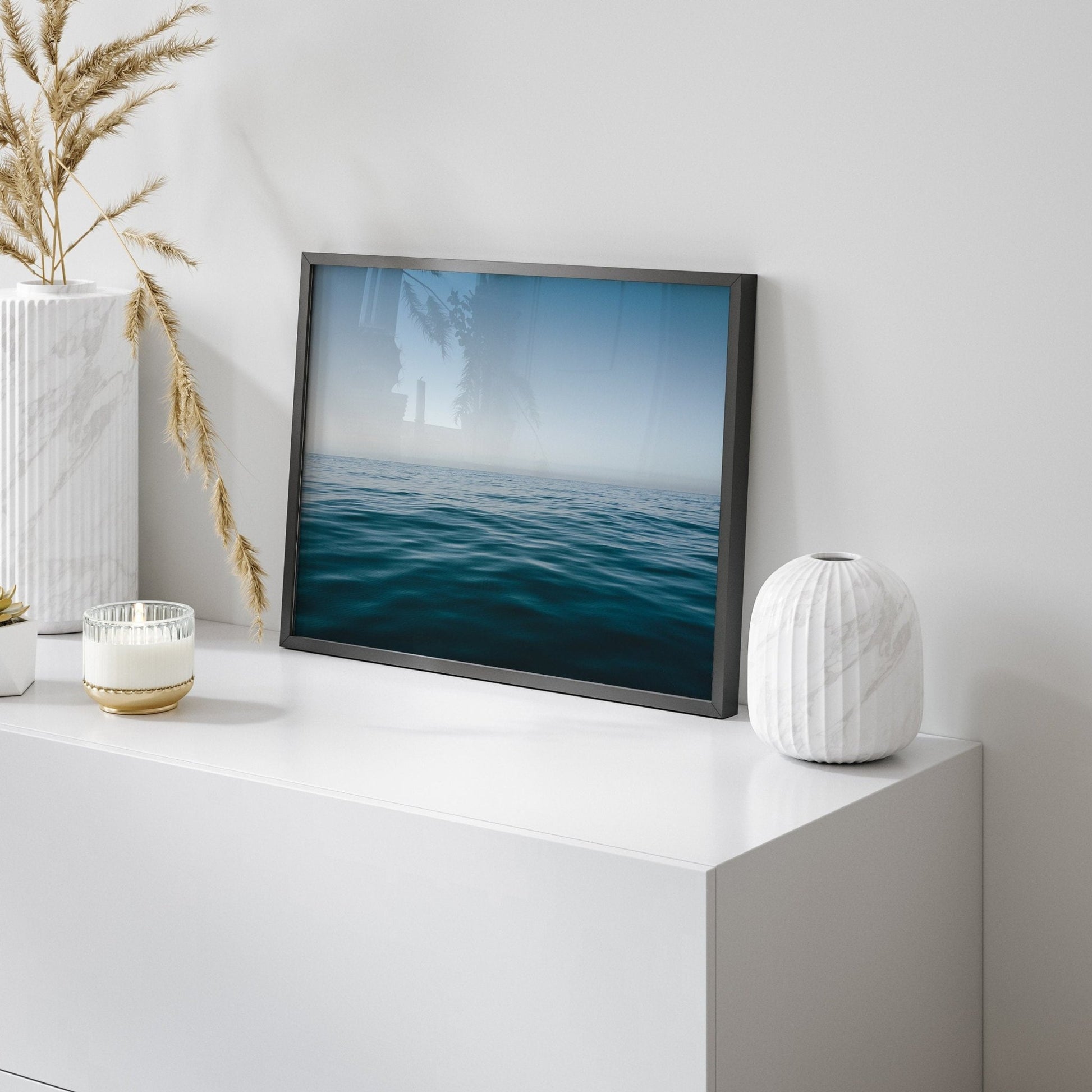 The Zen Ocean photo rests on a white table with a vase, candle, and dried plant.