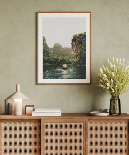 Framed photograph of Ninh Binh, Vietnam in a neutral frame against light green walls. Photograph features a small boat in the waterway between tall limestones